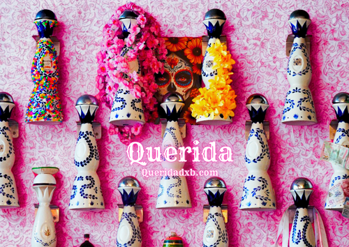 Decorative figurines with colorful attire on a pink background, featuring the brand 'Querida'.