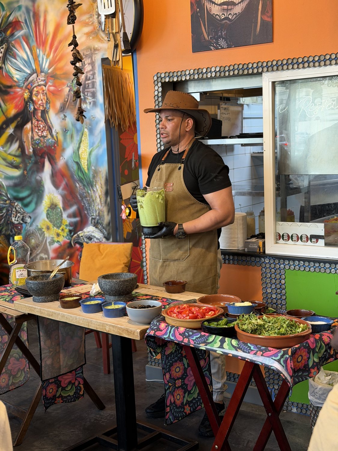 Chef Ernesto hosting a Querida masterclass in his straw hat and apron, blending fresh salsa at the demo table