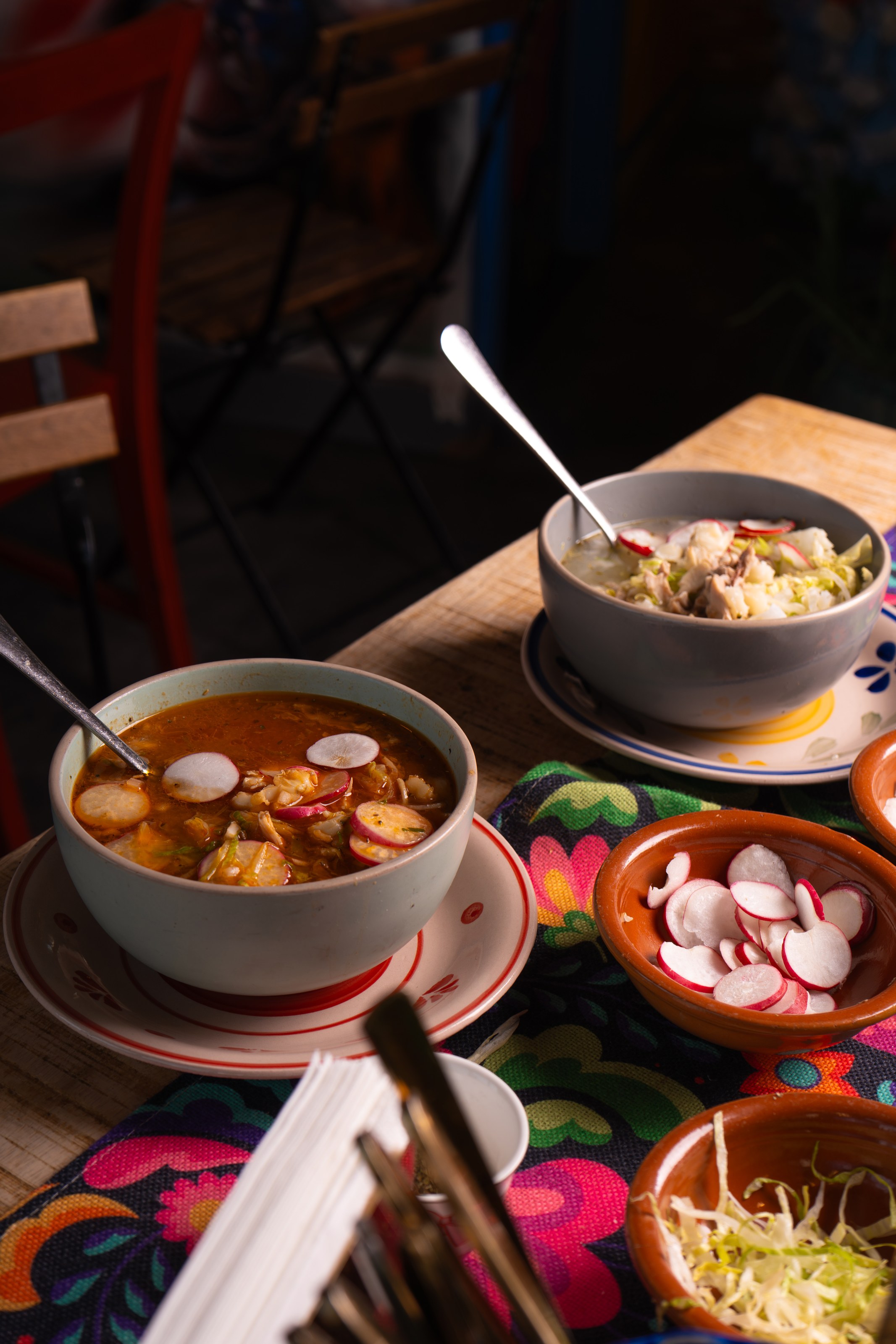 Pozole rojo and pozole blanco side by side, deep side-light, on the family table