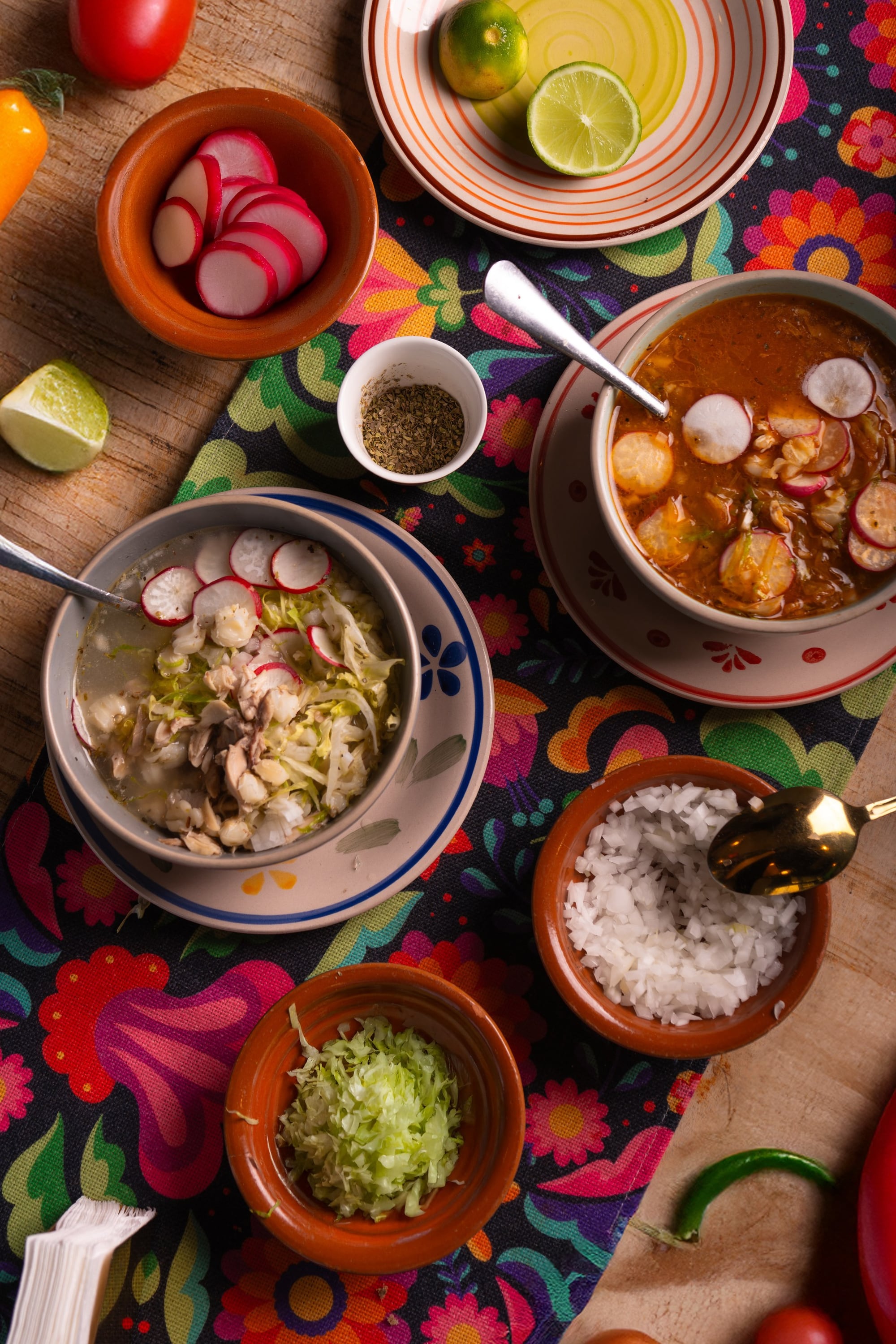 Top-down spread — both pozoles, radish, cabbage, lime, oregano, rice, on the floral runner