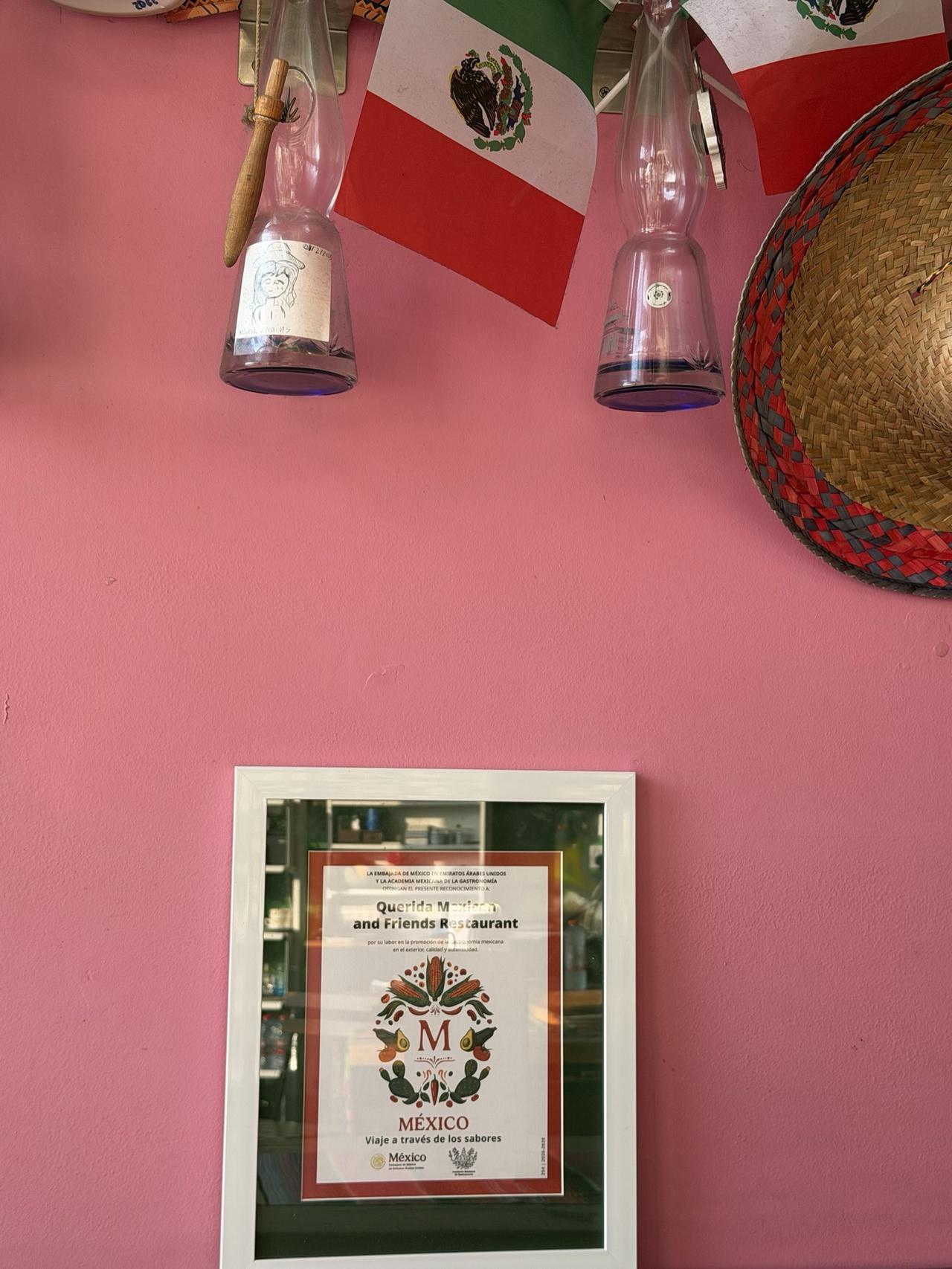 The framed certificate on Querida's pink wall, below two small Mexican flags, two glass bottles and a straw sombrero