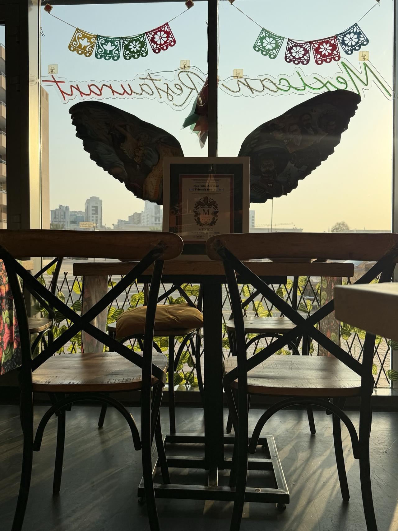 The framed Reconocimiento No. 254 standing on a table in Querida's front window, between the two painted wings, with papel picado above and Al Barsha 1 outside at sunset