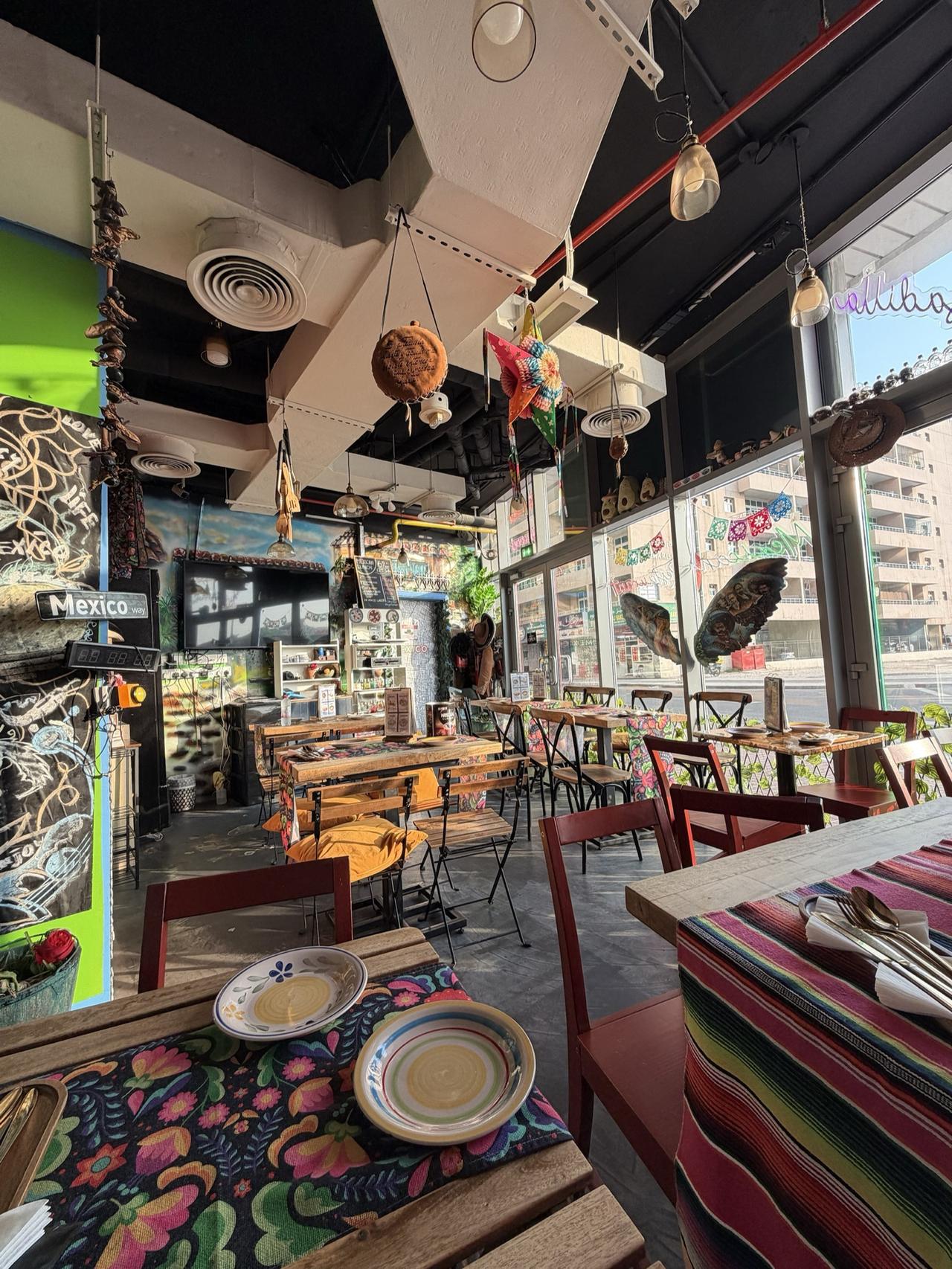 Querida's 26-seat dining room: wooden tables with floral and striped runners, red chairs, a piñata star hanging from the ceiling, and the painted wings in the window with 14th Street outside